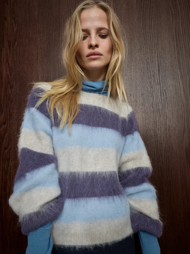 A woman wearing a fuzzy, oversized striped sweater in light blue, lavender, and cream, layered over a light blue turtleneck, standing against a dark wood-paneled wall.