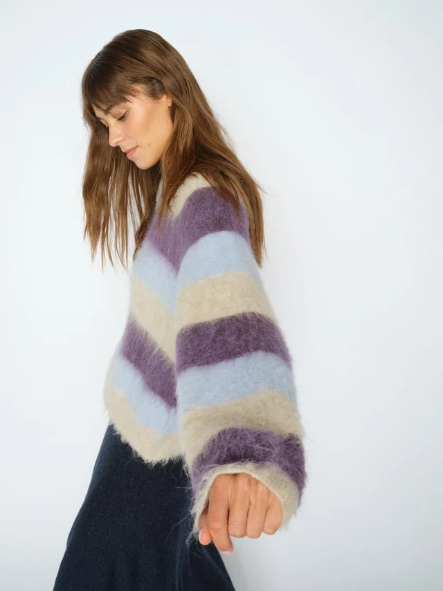 A woman wearing a fuzzy, oversized striped sweater with horizontal bands of purple, light blue, and cream, paired with a dark skirt, photographed against a plain white studio background.