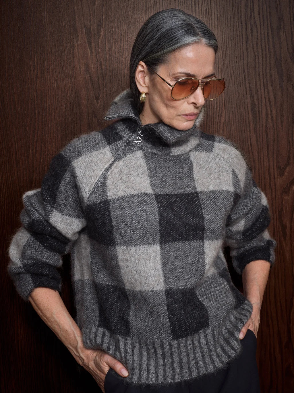A woman wearing a gray and black buffalo plaid zip-up sweater, styled with brown aviator sunglasses, standing against a dark wood-paneled background.