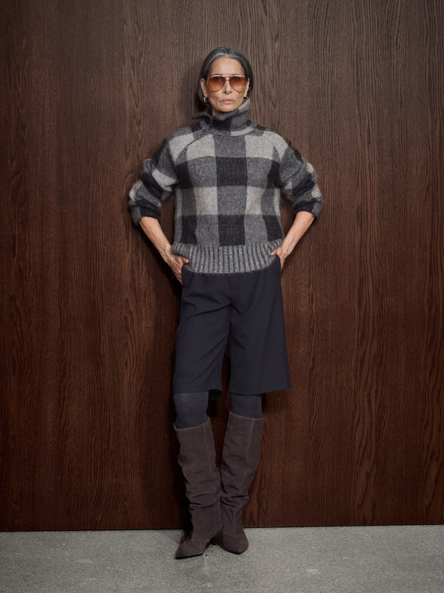 A woman wearing a gray and black buffalo plaid zip-up sweater, styled with brown aviator sunglasses, standing against a dark wood-paneled background.
