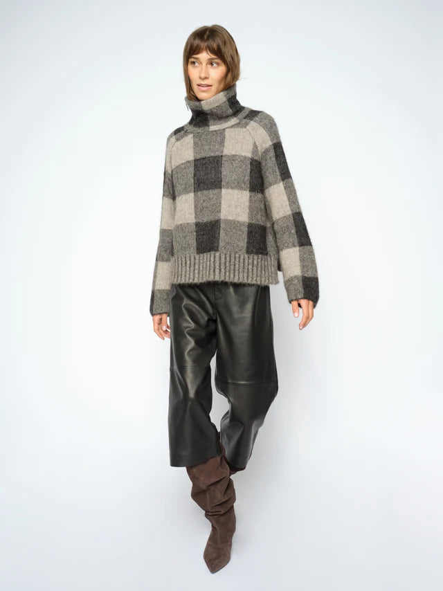 A model wearing a gray and black checkered knit turtleneck sweater, black leather pants, and brown slouchy boots, standing against a light gray studio background.