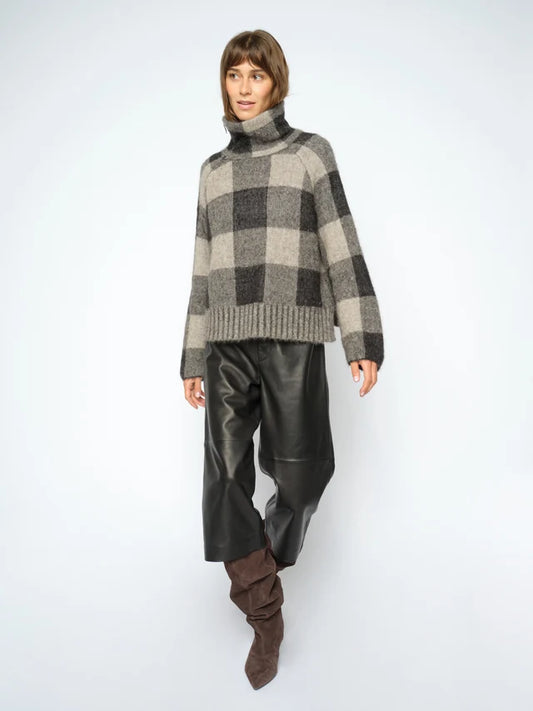 A model wearing a gray and black checkered knit turtleneck sweater, black leather pants, and brown slouchy boots, standing against a light gray studio background.
