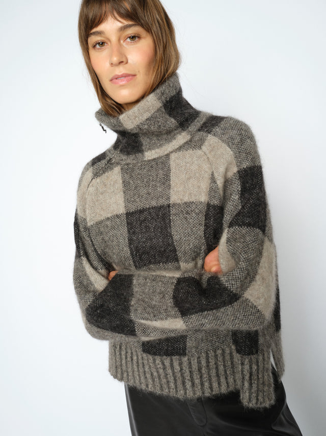A woman wearing a fuzzy high-neck turtleneck sweater with a large-scale black, gray, and beige plaid pattern, posed against a light gray studio background.