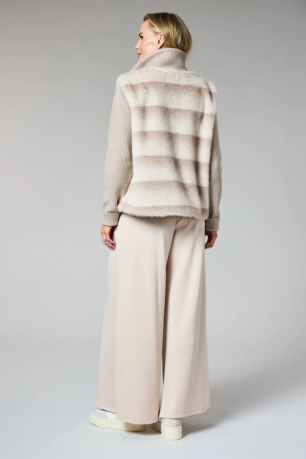 A model shown from the back wearing a beige and taupe striped faux fur vest over a beige knit sweater and wide-leg beige trousers, set against a plain light gray studio background.