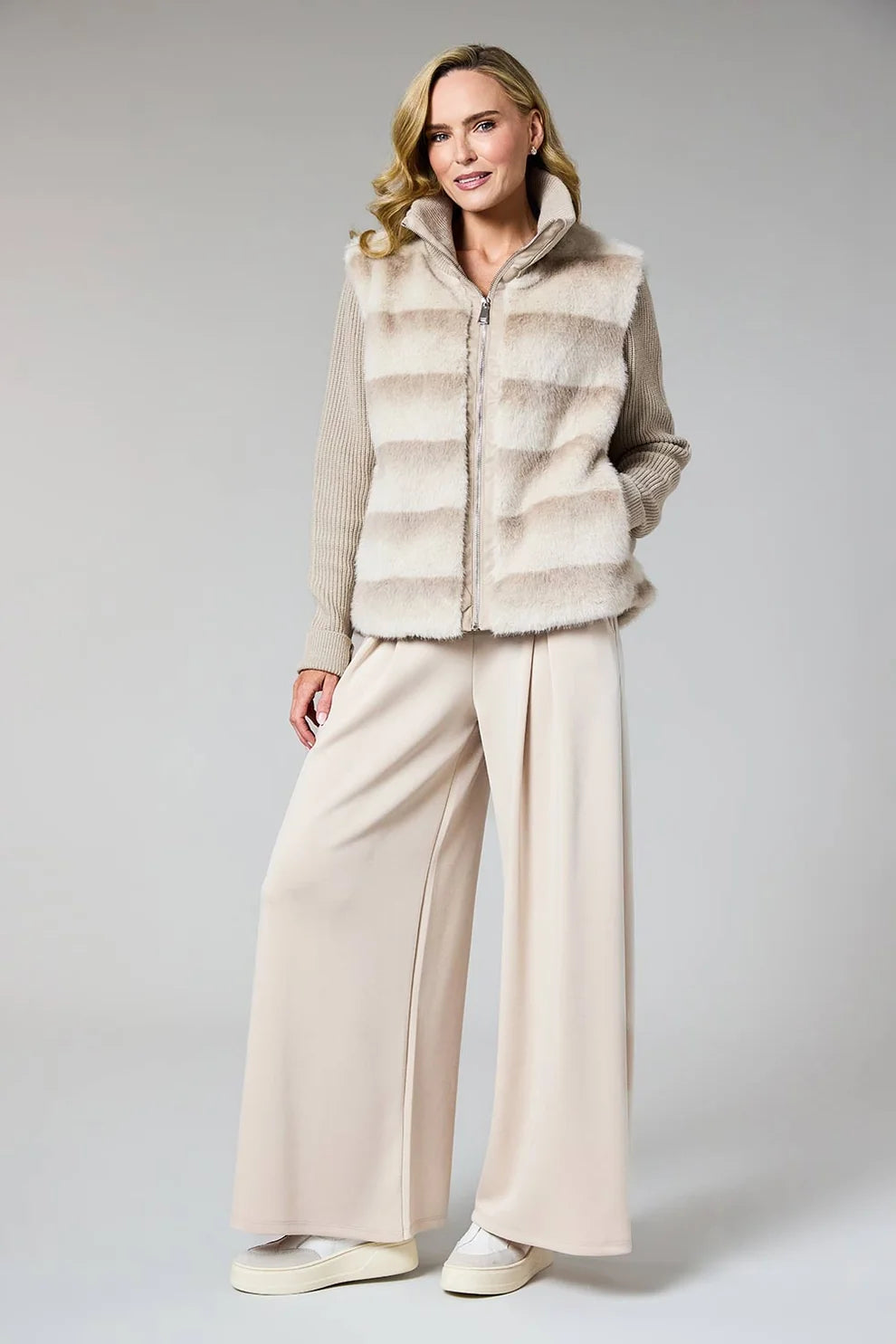 A woman modeling a beige faux-fur zip-front vest over a beige long-sleeve top and wide-leg beige trousers, standing against a light gray studio background.