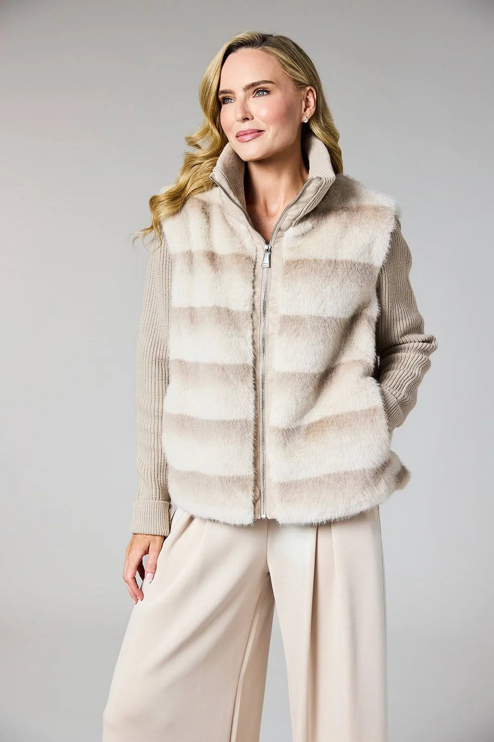 A woman modeling a beige and cream faux fur vest over a ribbed knit sweater and cream wide-leg trousers, photographed against a neutral light gray studio background.