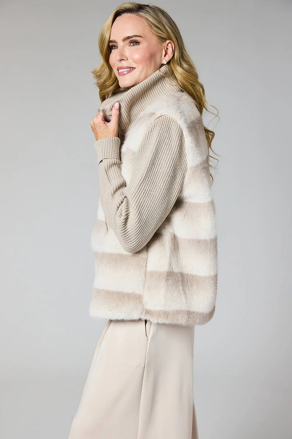 A woman wearing a beige and cream striped faux fur vest over a ribbed knit turtleneck sweater and matching beige trousers, photographed against a neutral light gray studio background.