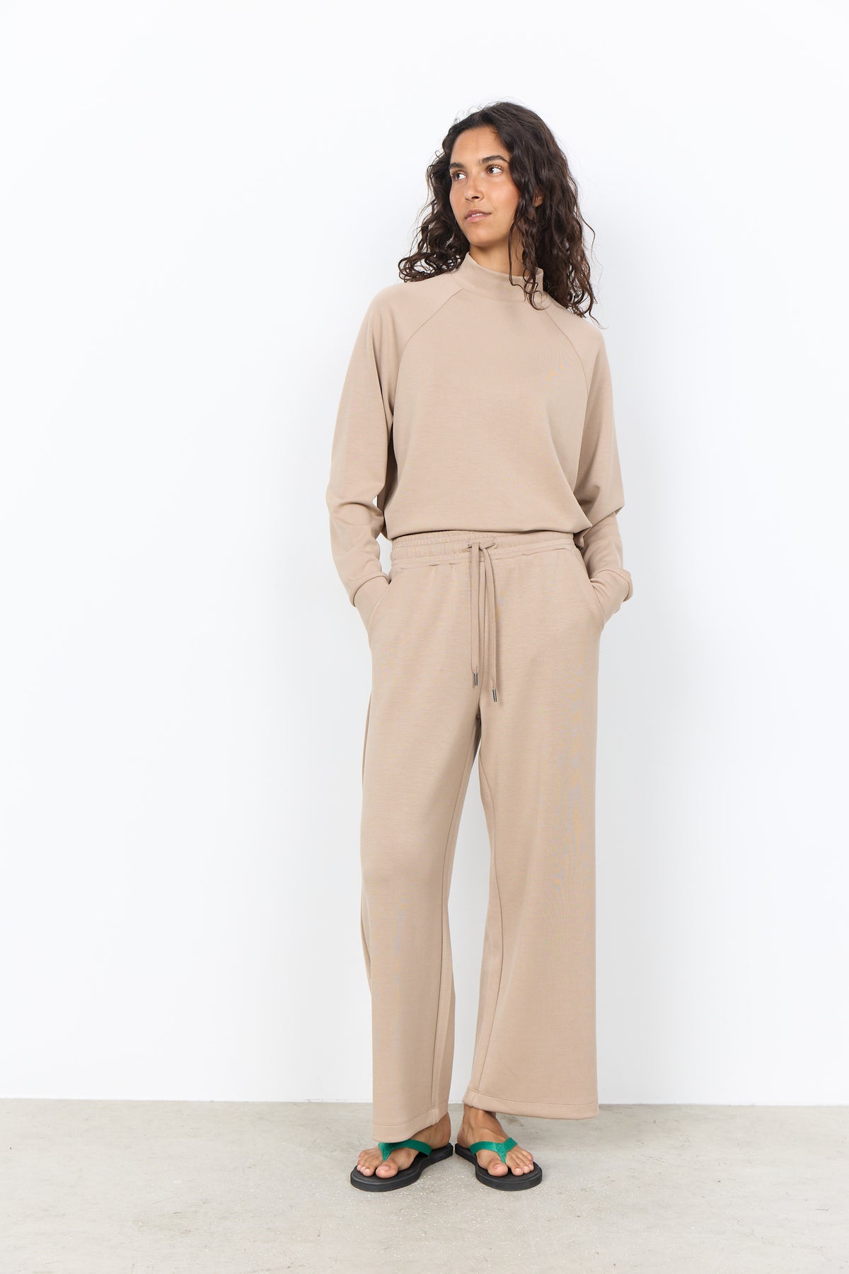 A woman modeling a matching beige knit lounge set consisting of a long-sleeve mock-neck top and wide-leg drawstring pants, paired with green sandals, against a plain white studio background.
