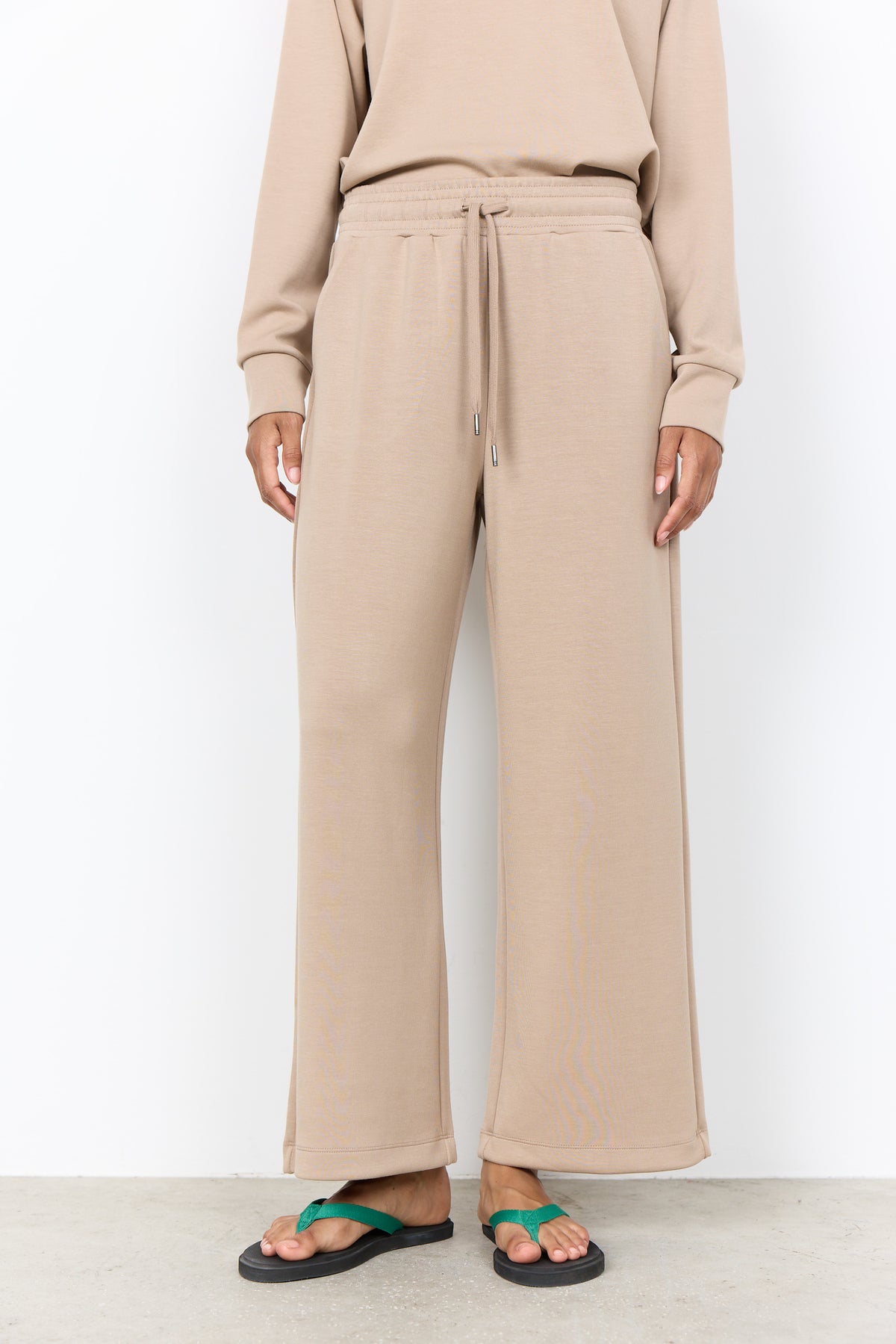 A person wearing beige wide-leg drawstring sweatpants with a matching sweatshirt and green flip-flop sandals, standing against a plain white wall.