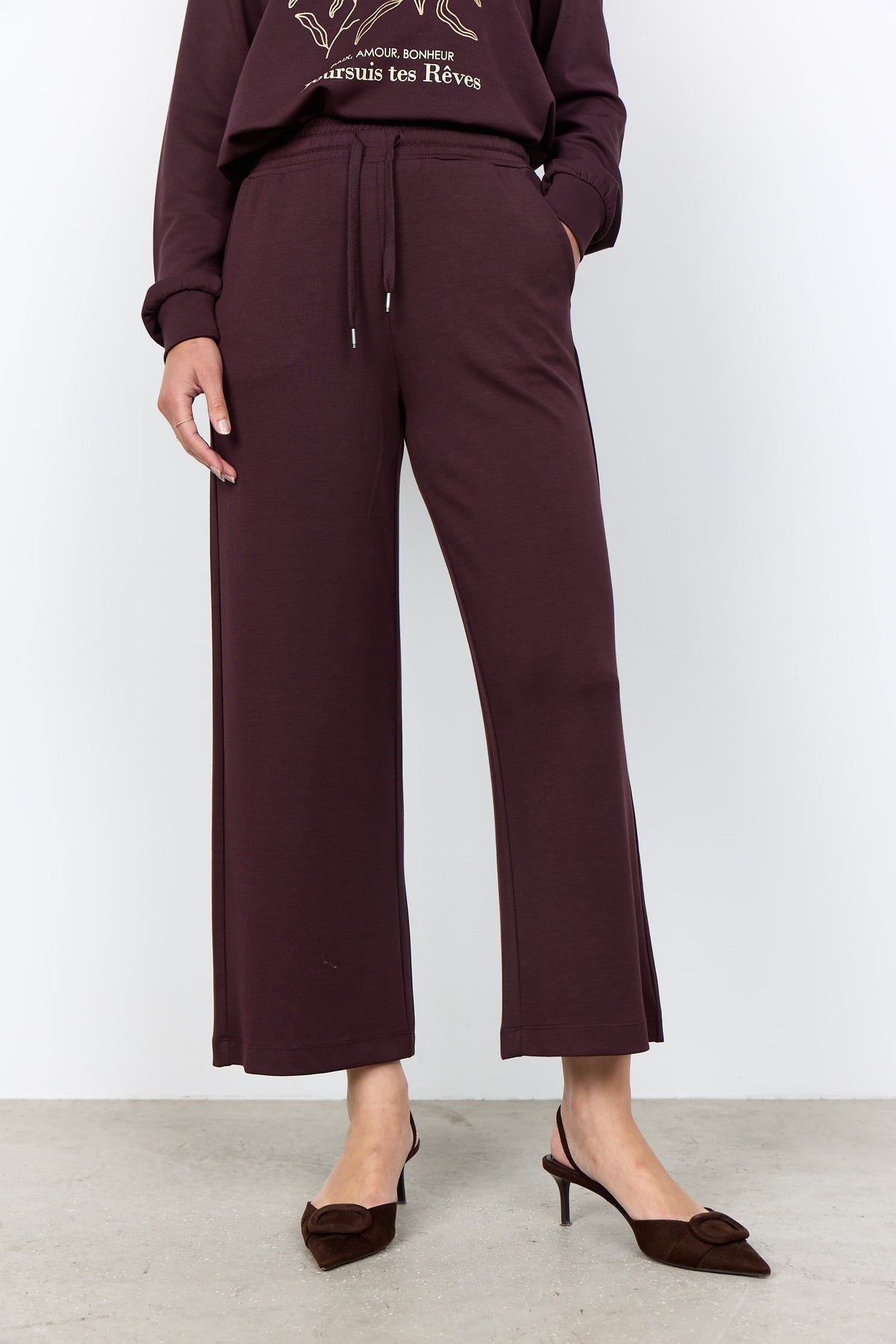 Person wearing a dark purple outfit with wide-leg pants and a cropped top against a white background