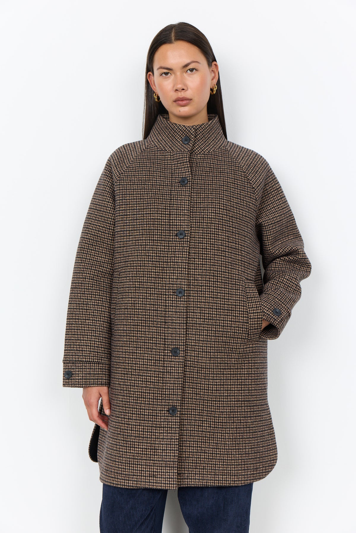 A woman modeling a brown and beige houndstooth patterned coat with a high stand collar and front buttons, paired with dark blue jeans, against a plain white background.