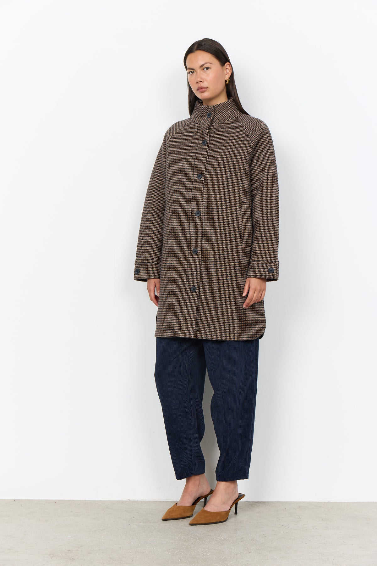 A model stands against a plain white studio wall wearing a brown and black checkered coat, dark navy pants, and tan pointed-toe heels.