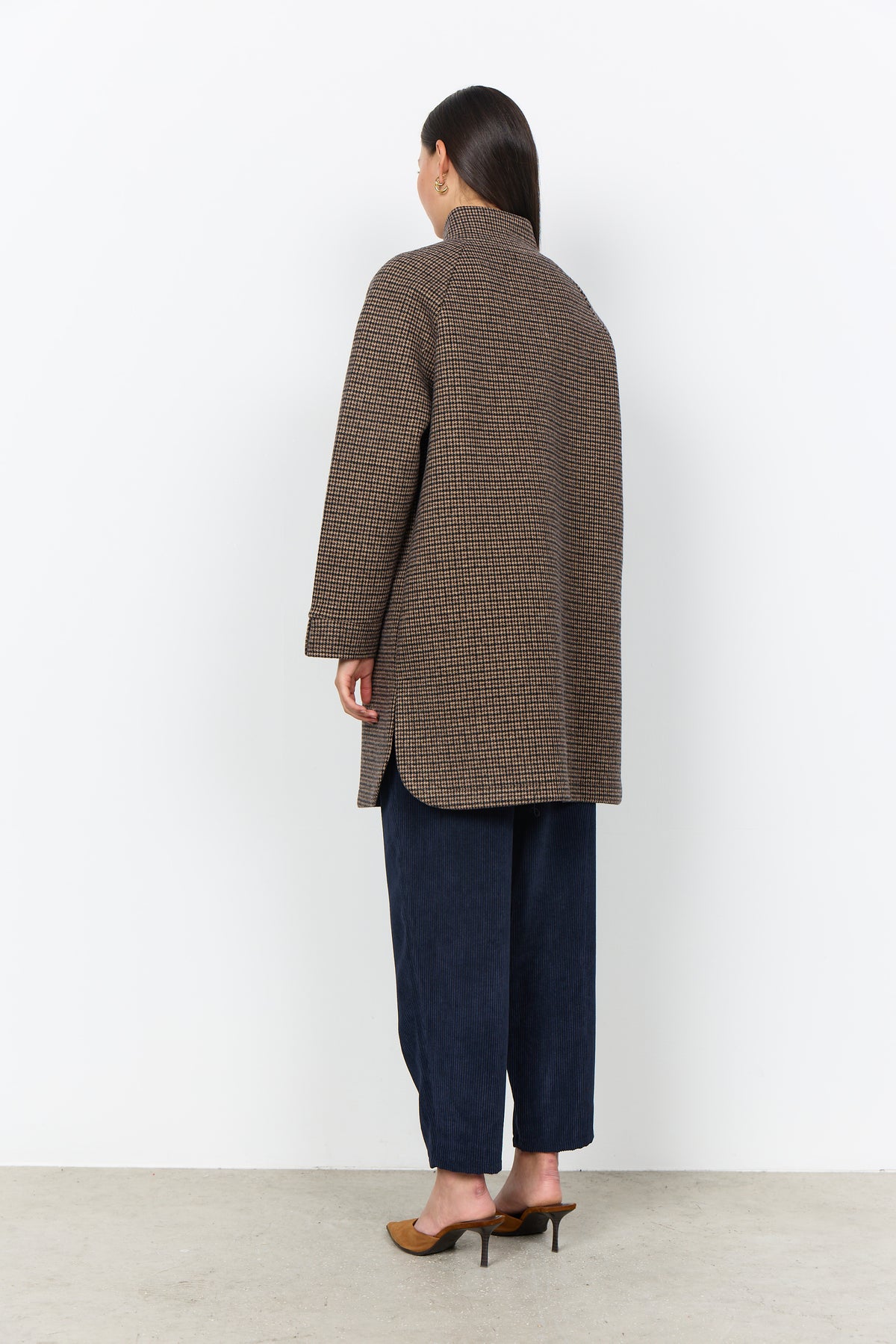 A model shown from the back wearing a brown and black houndstooth patterned coat, dark blue wide-leg trousers, and brown pointed-toe heels against a plain white wall.