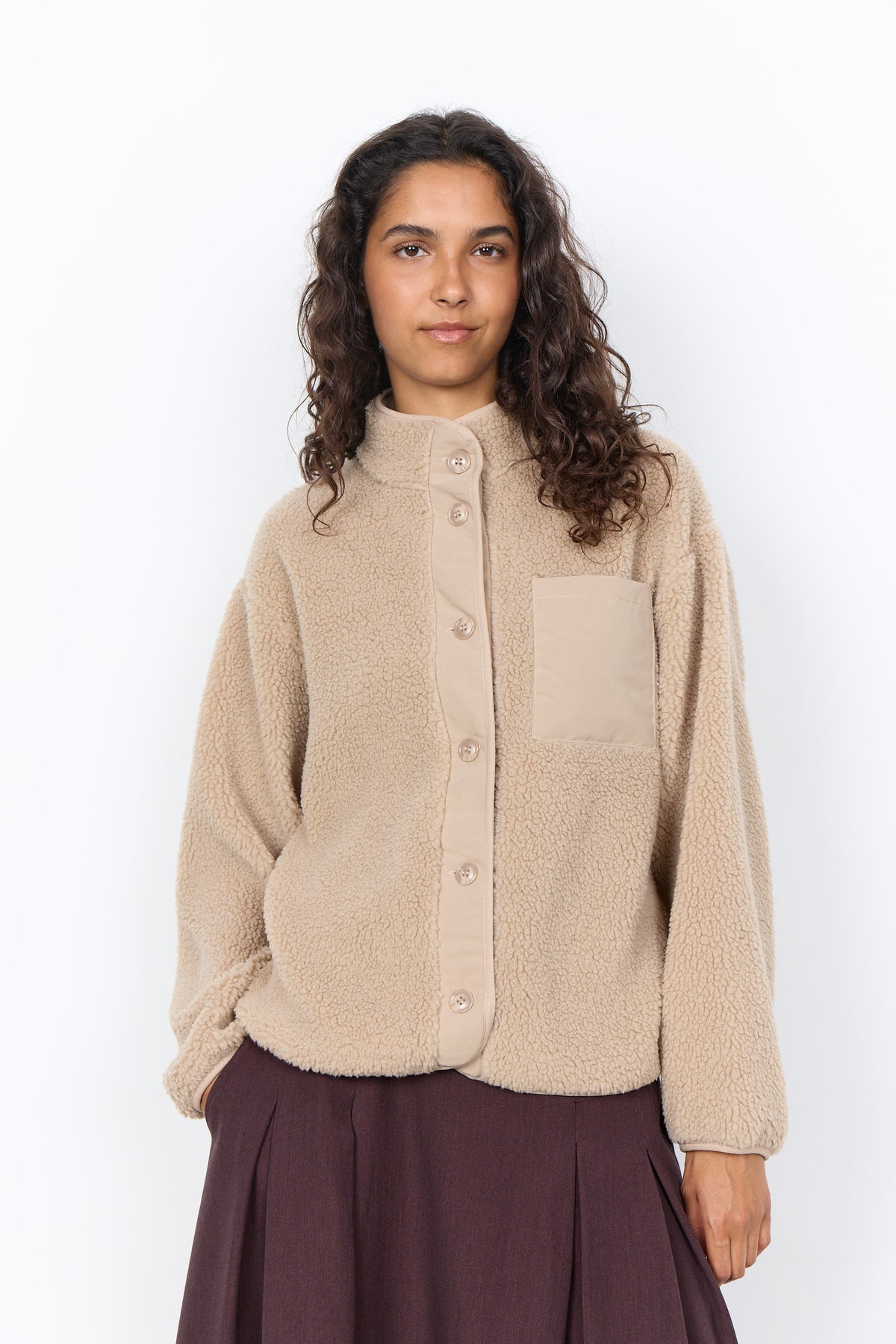 A woman with curly brown hair wearing a beige sherpa fleece button-up cardigan with a chest pocket, paired with a dark purple skirt, against a plain white background.