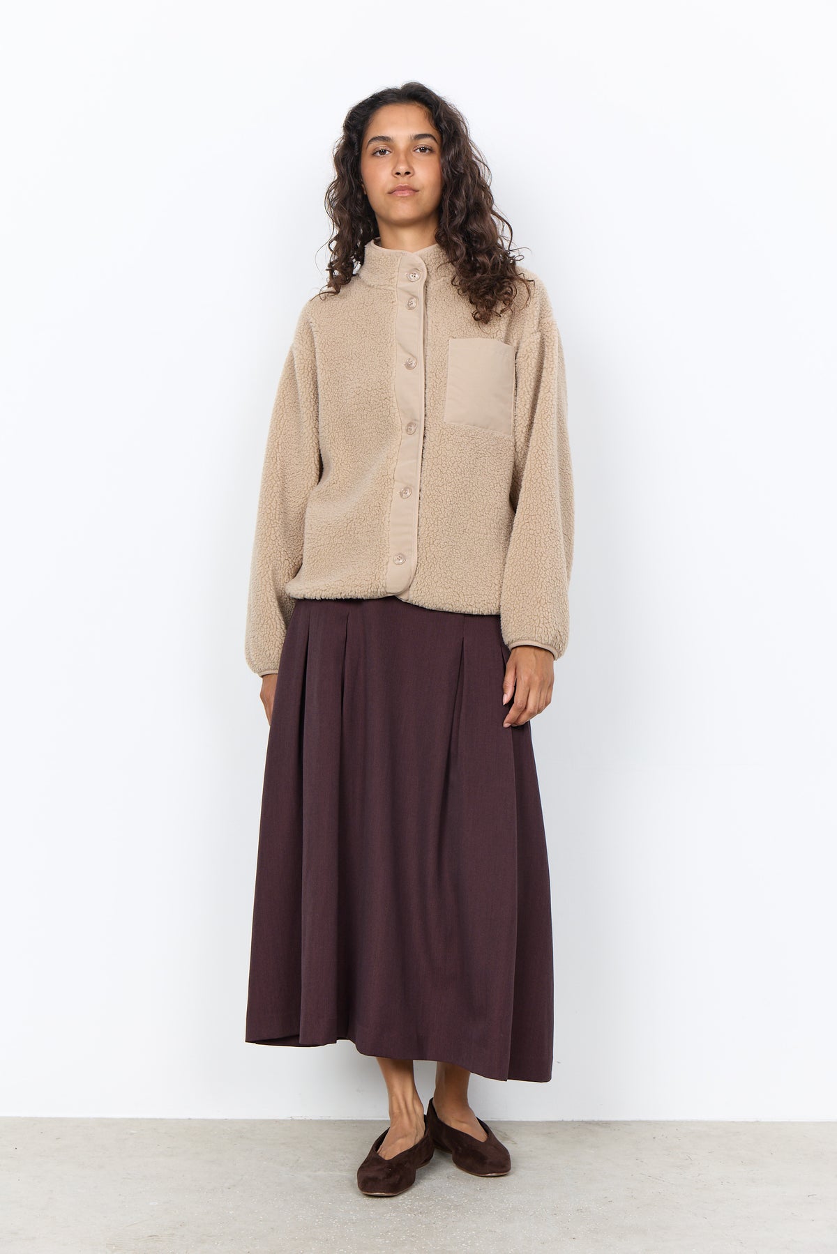 A woman stands against a plain white wall wearing a beige cropped button-front cardigan with a chest pocket and a dark plum midi skirt, paired with brown flats.