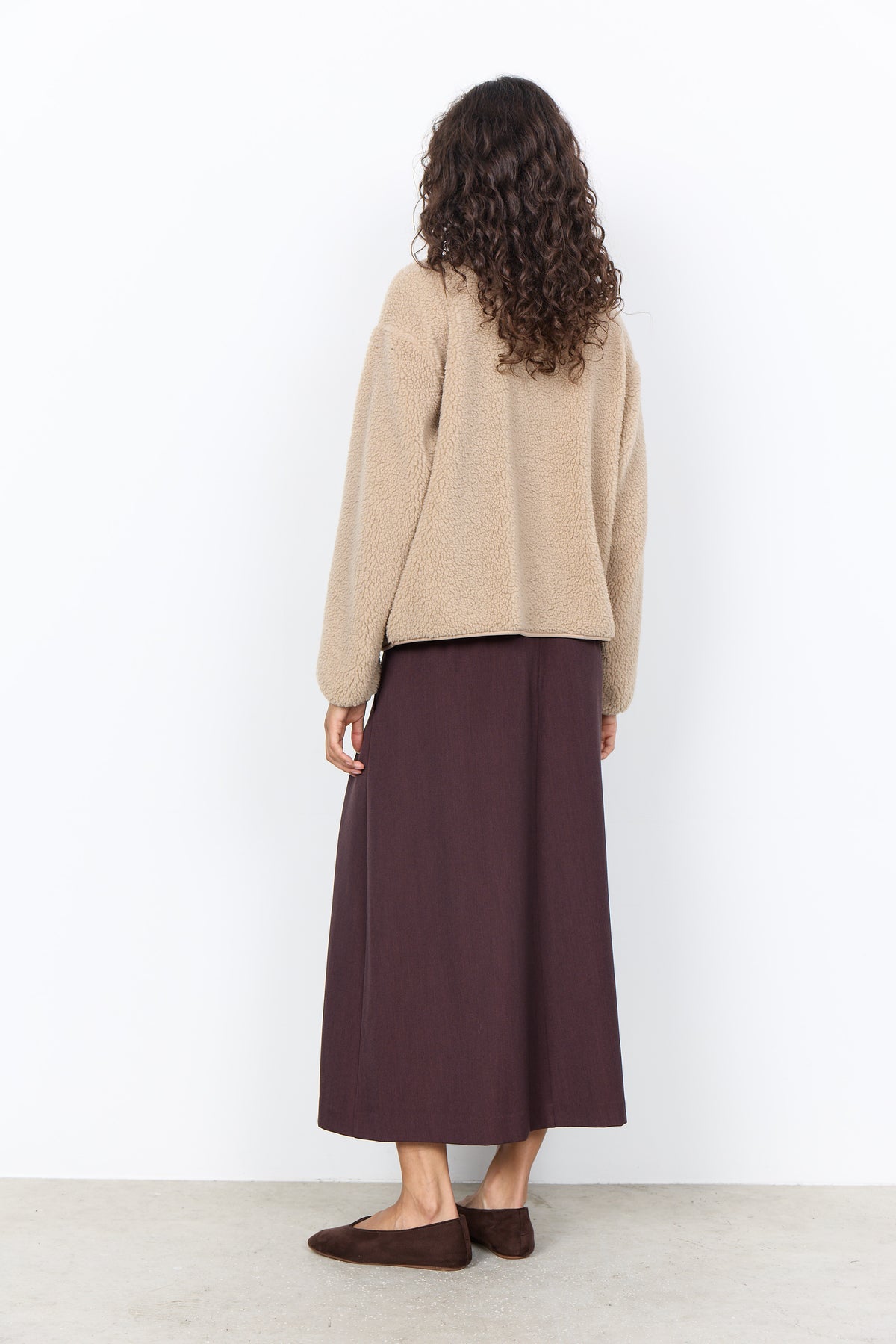 A woman shown from the back wearing a beige knit cardigan and a dark plum midi skirt, paired with brown flat shoes, against a plain white wall.