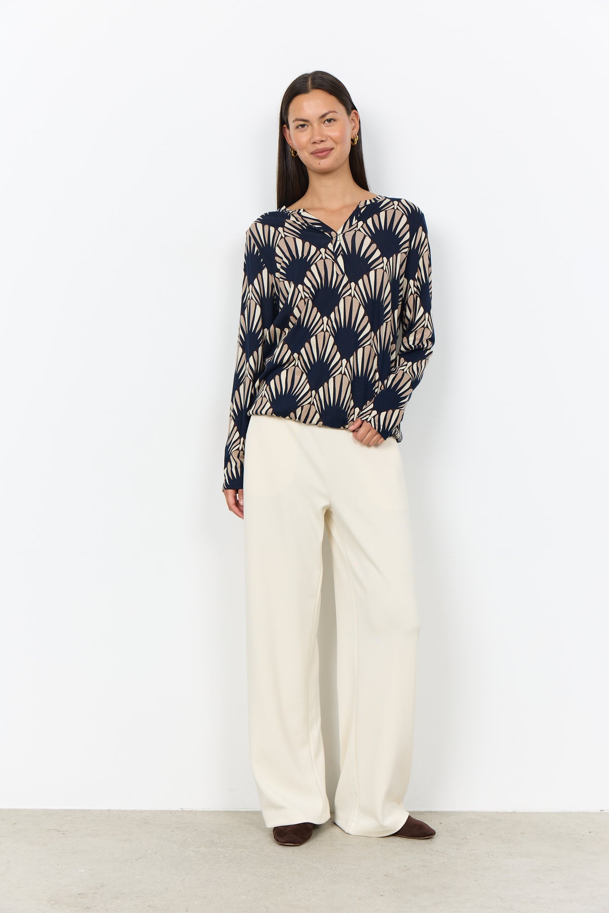 A woman modeling a long-sleeve knit top with a navy and beige fan-like geometric pattern, paired with wide-leg cream trousers, standing against a plain white wall.