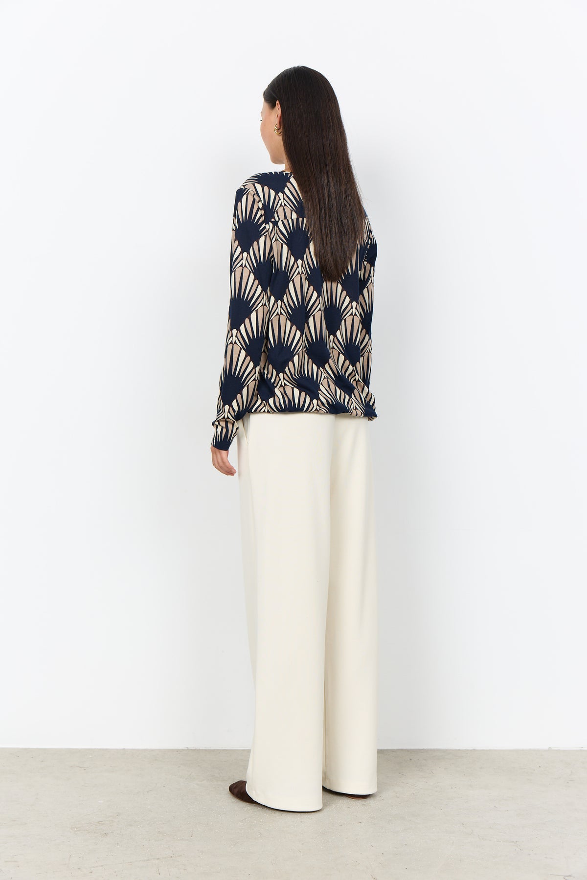 A woman is shown from the back wearing a navy and beige geometric patterned long-sleeve top and cream wide-leg trousers, standing against a plain white wall.