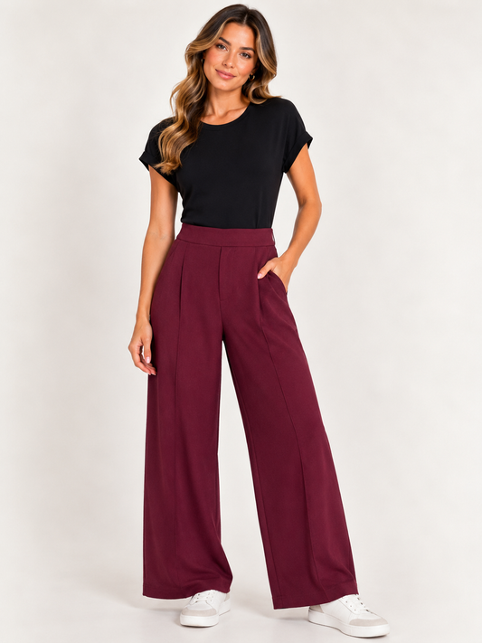 A woman modeling a pair of high-waisted, wide-leg pleated trousers in burgundy, paired with a black t-shirt and white sneakers, against a plain light background.