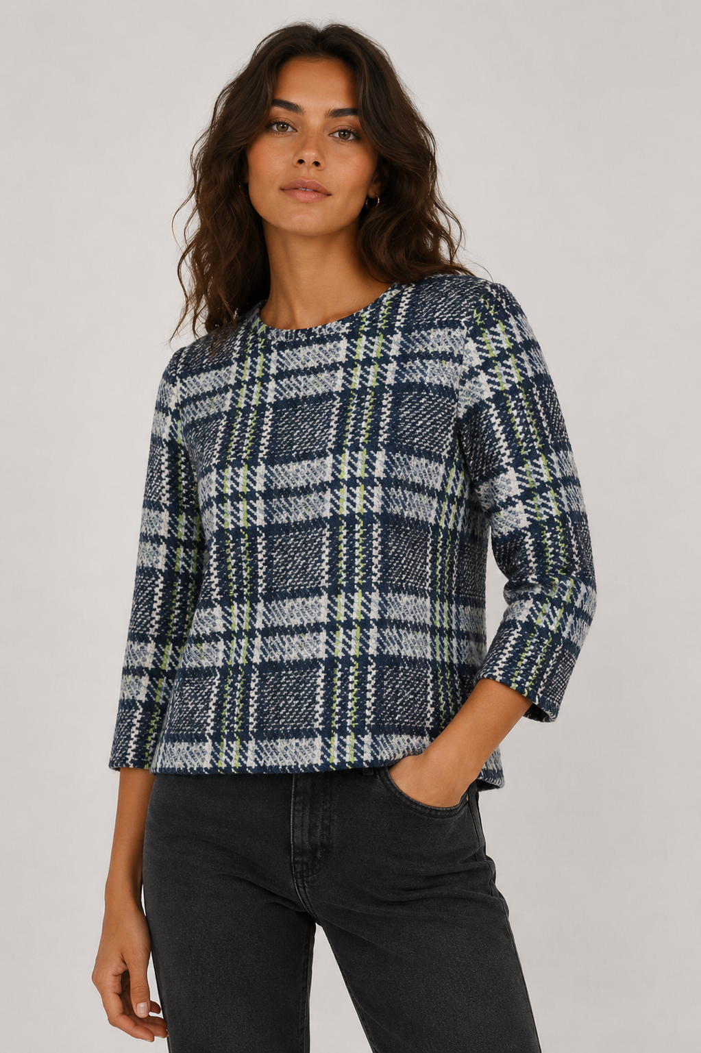 A woman wearing a textured plaid knit top with three-quarter sleeves and dark gray jeans, standing against a light gray studio background.