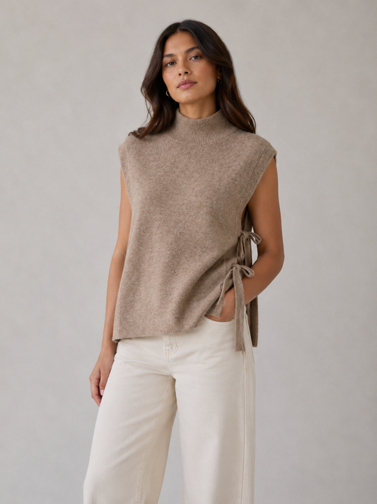 A woman modeling a sleeveless taupe knit mock neck top with side tie details, paired with off-white wide-leg pants, against a neutral gray studio background.