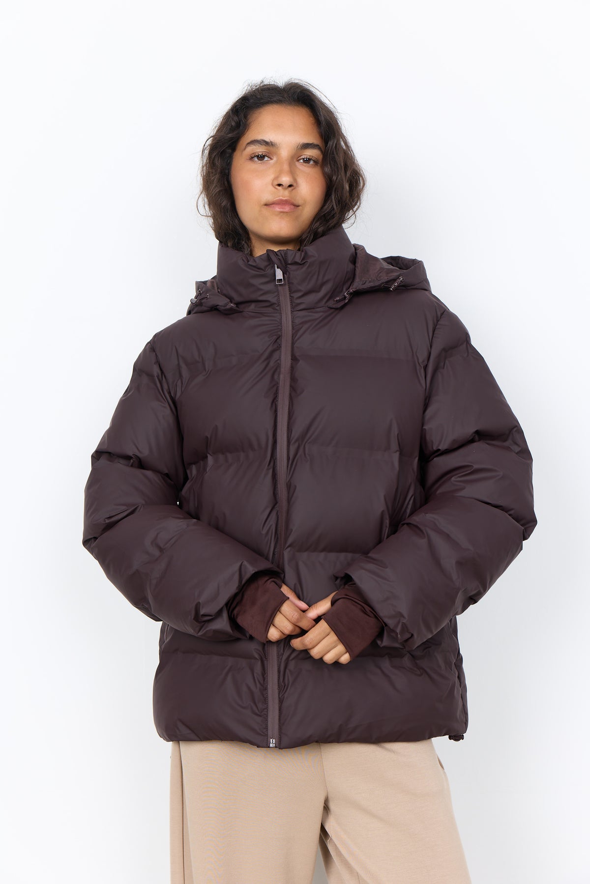 A model wearing a dark brown hooded puffer jacket with a high collar and front zipper, paired with beige pants, photographed against a plain white background.