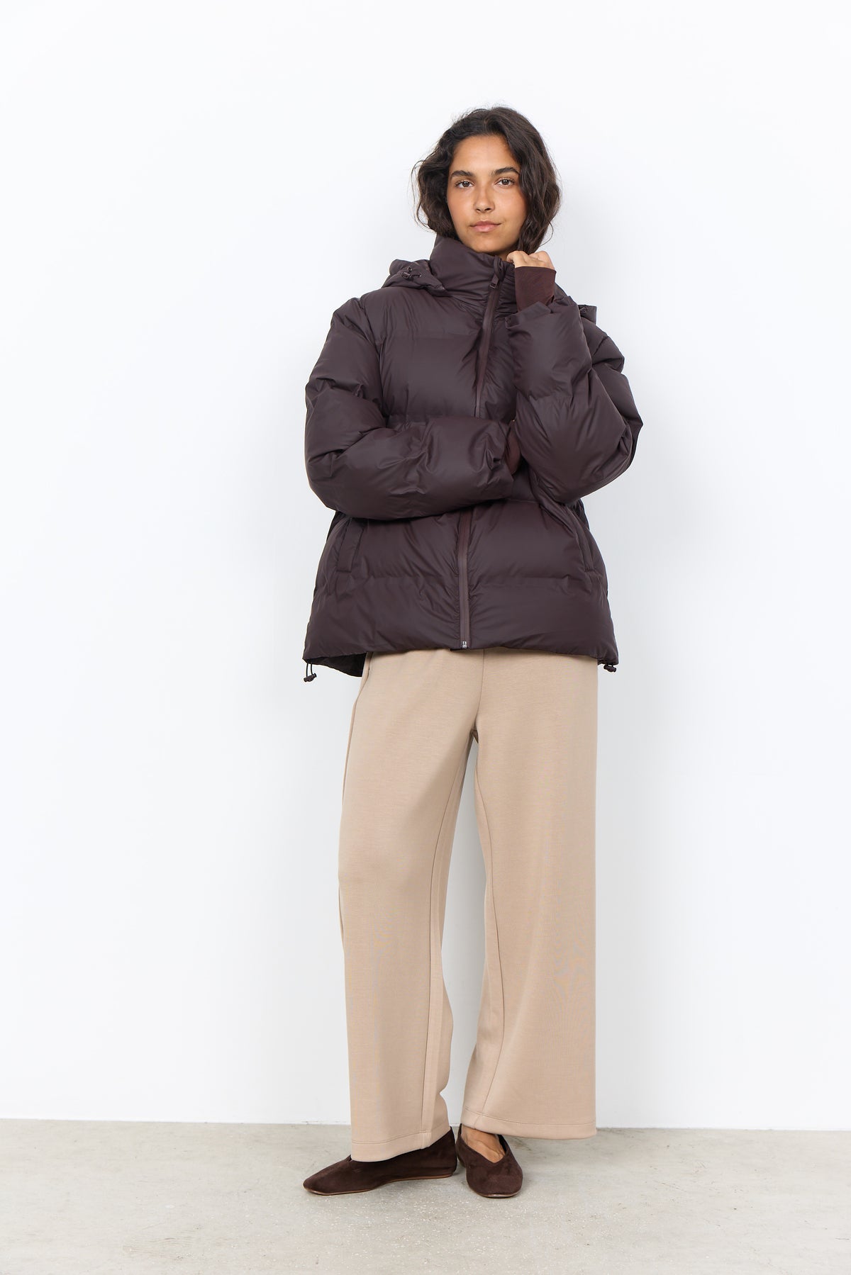 A model stands against a plain white studio wall wearing a dark brown puffer jacket, beige wide-leg trousers, and dark brown flat shoes.