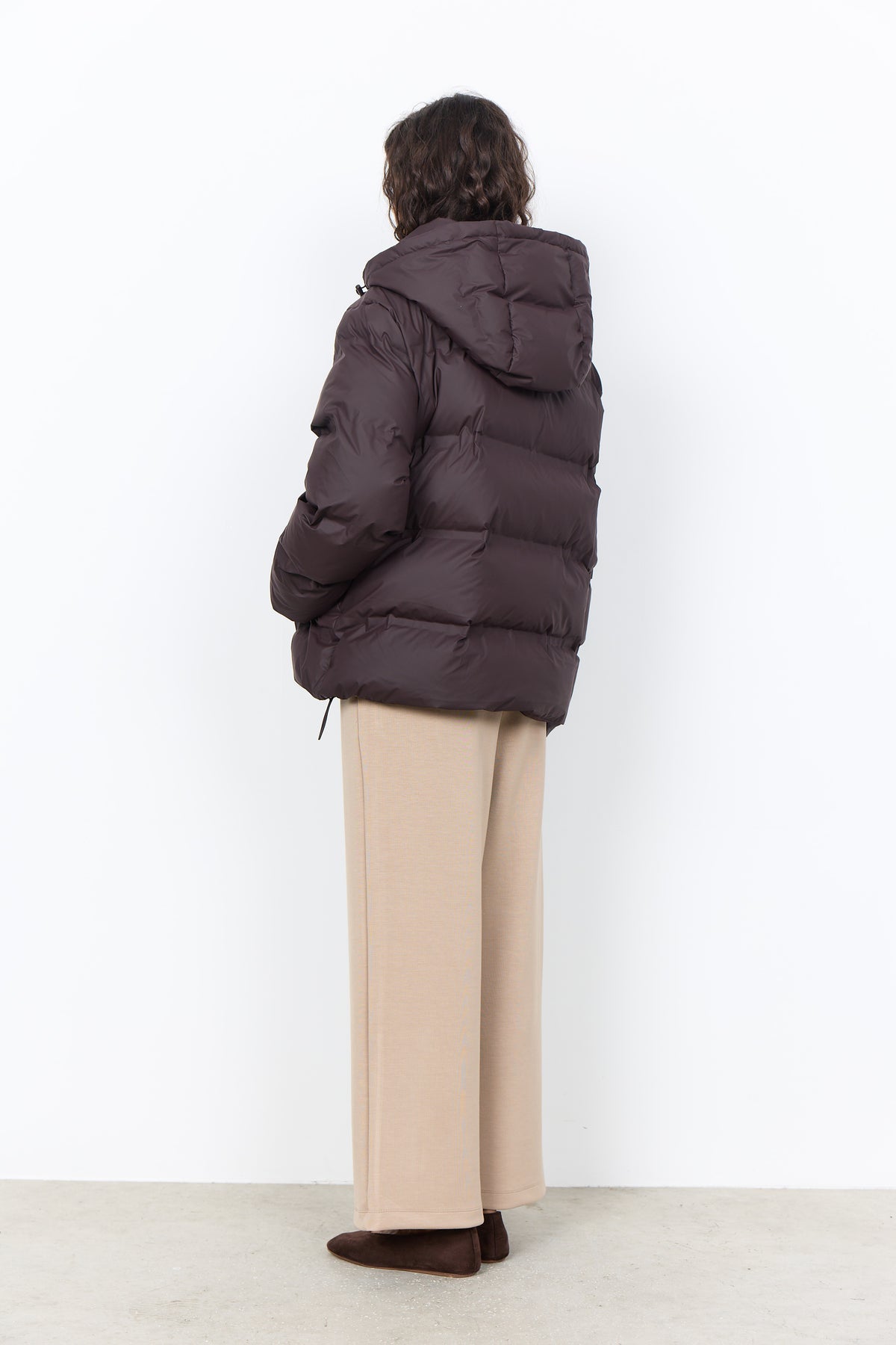 A model shown from the back wearing a dark brown puffer jacket, beige wide-leg trousers, and brown shoes, standing against a plain white wall.