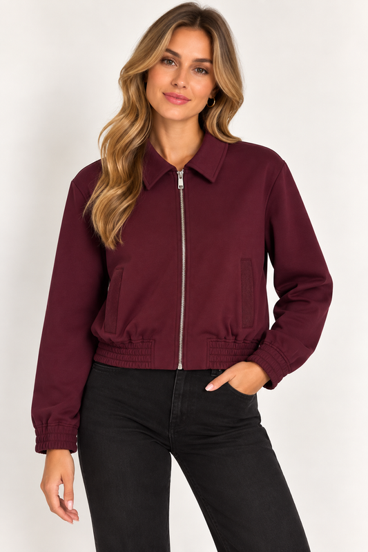 A woman wearing a cropped burgundy zip-up bomber jacket with black pants, standing against a plain light gray studio background.