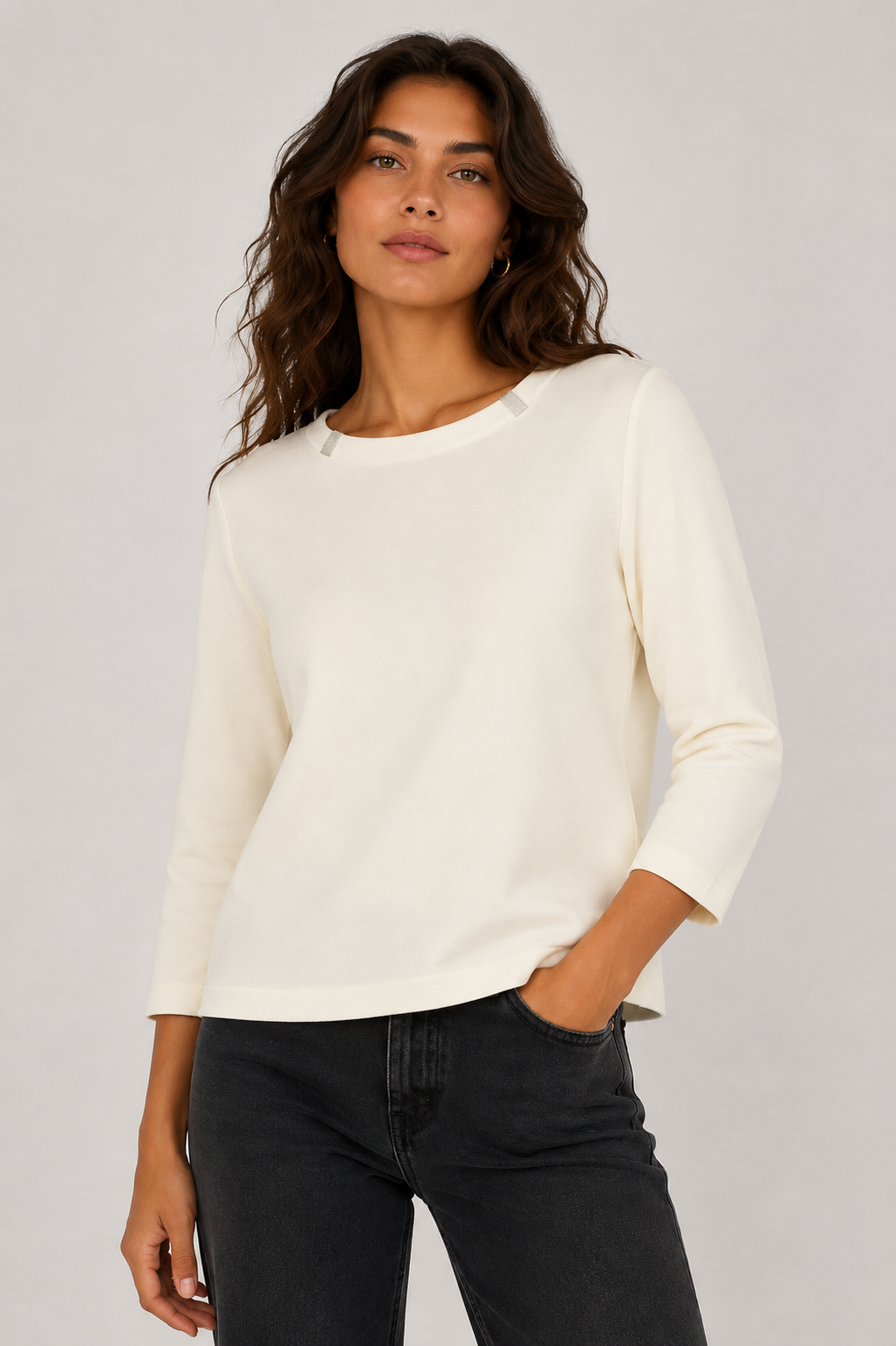 A woman wearing a cream-colored three-quarter sleeve top and black jeans, standing against a light gray studio background.