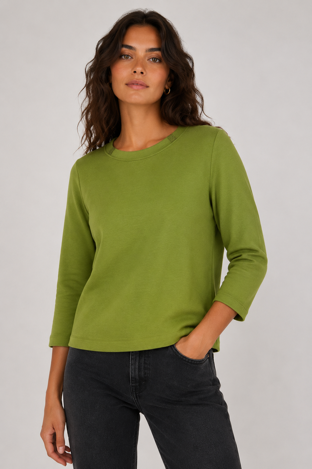 A woman wearing a sage green three-quarter sleeve top and dark gray jeans, standing against a light gray studio background.