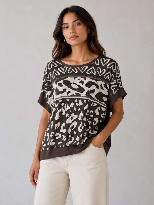 A woman modeling a short-sleeve knit top with a mixed pattern design featuring a leopard print on the lower half, a heart pattern on the upper chest, and solid dark brown trim, paired with cream pants against a light gray studio background.