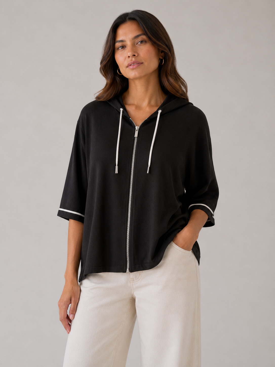 A woman wearing a black zip-up hoodie with white drawstrings and white piping on the sleeves, paired with off-white trousers, against a neutral gray studio background.