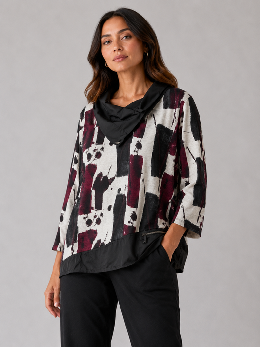 A woman modeling a black, white, and burgundy abstract print top with a draped black collar, paired with black pants, against a solid gray studio background.