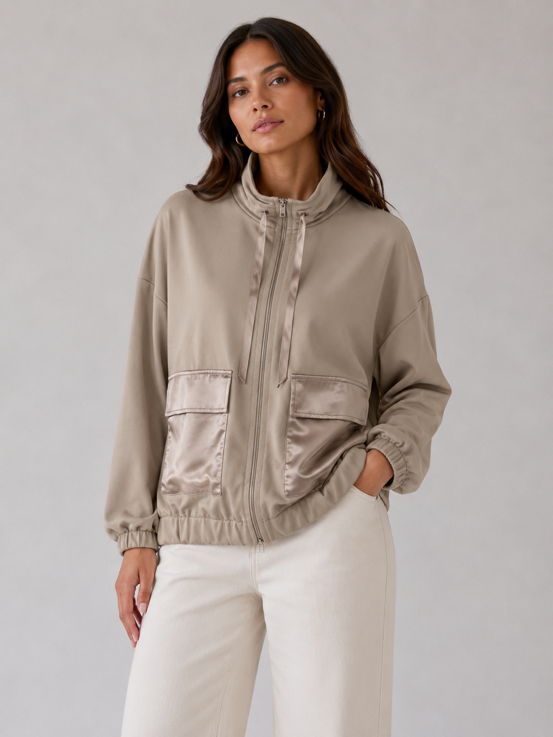 A woman modeling a cropped satin zip-up jacket in a light taupe-beige color, paired with off-white trousers, against a neutral gray studio background.