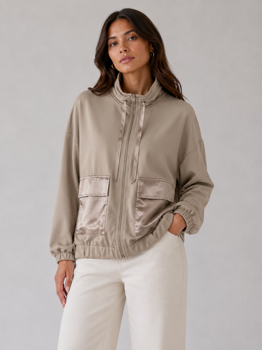 A woman modeling a cropped satin zip-up jacket in a light taupe-beige color, paired with off-white trousers, against a neutral gray studio background.