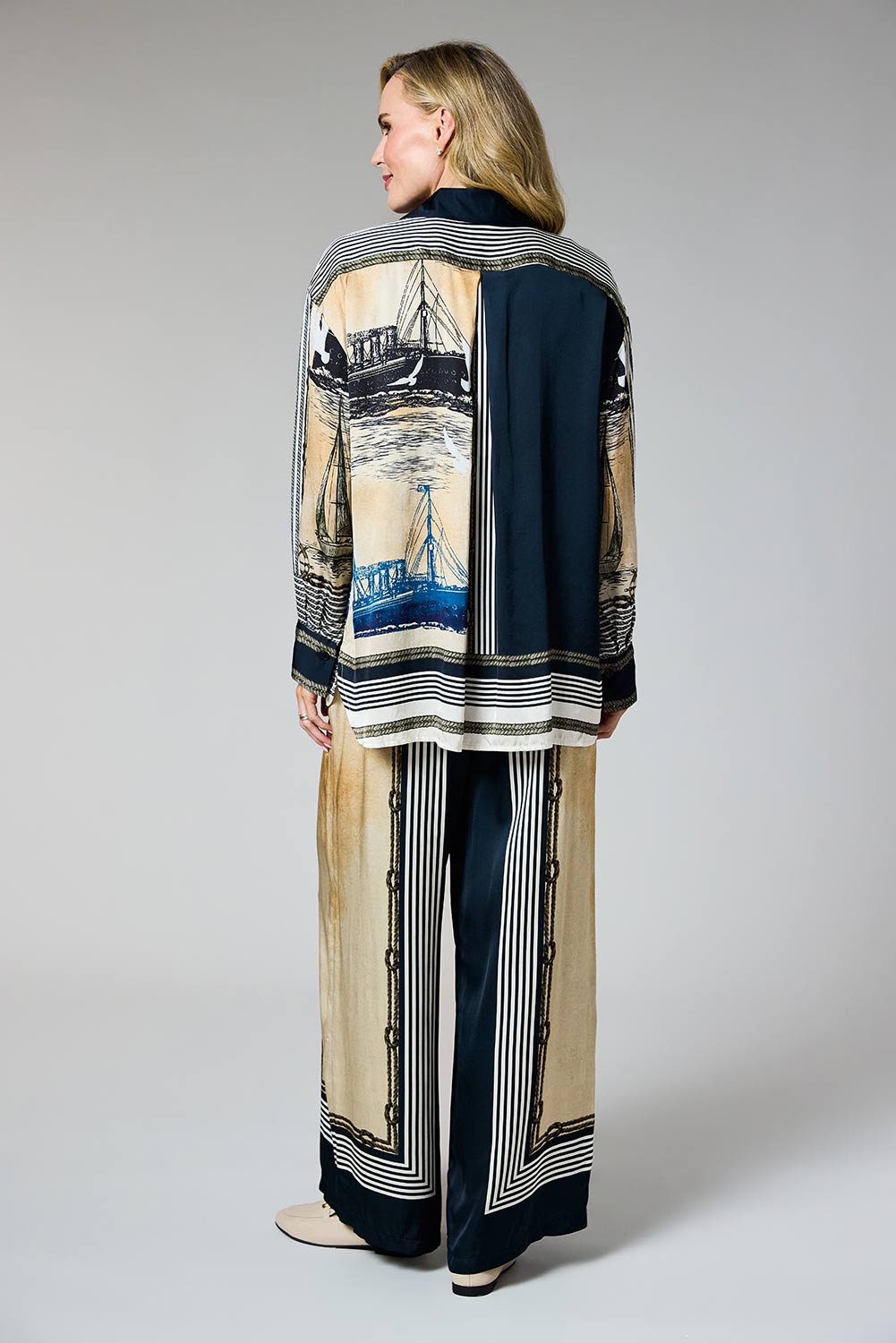 A woman stands with her back to the camera wearing a matching two-piece set consisting of a long-sleeve cardigan and wide-leg trousers. The set features a split design with one side in beige and blue nautical ship print and the other side in solid navy blue, with striped borders.