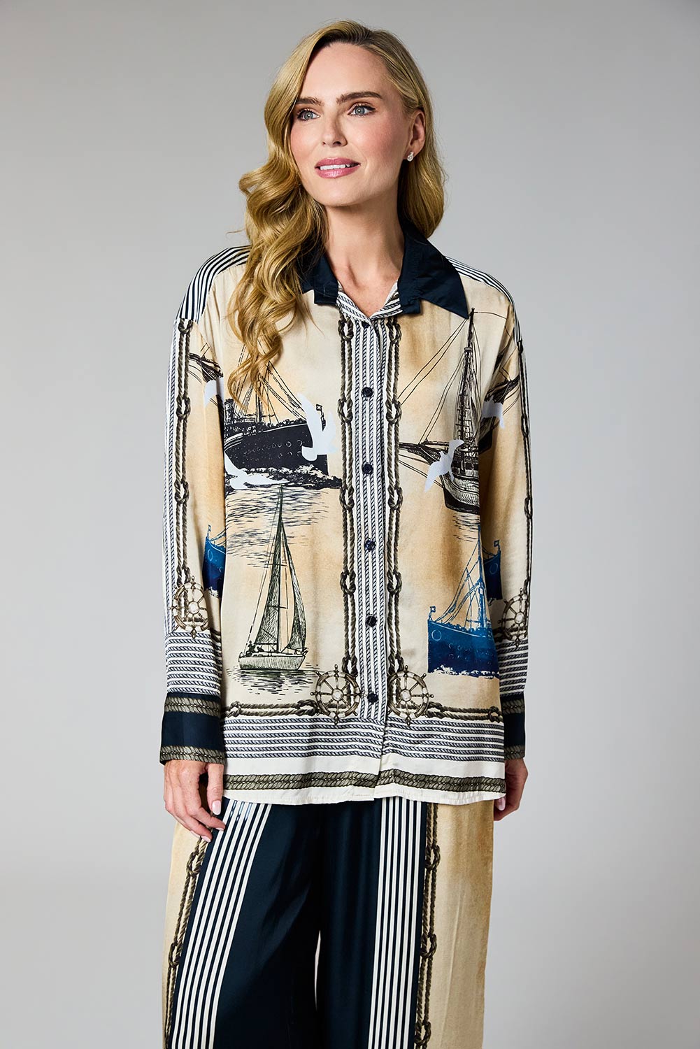 A woman modeling a long-sleeve button-front shirt with a beige and black nautical print featuring sailboats and chain motifs, paired with dark striped pants, against a neutral gray studio background.