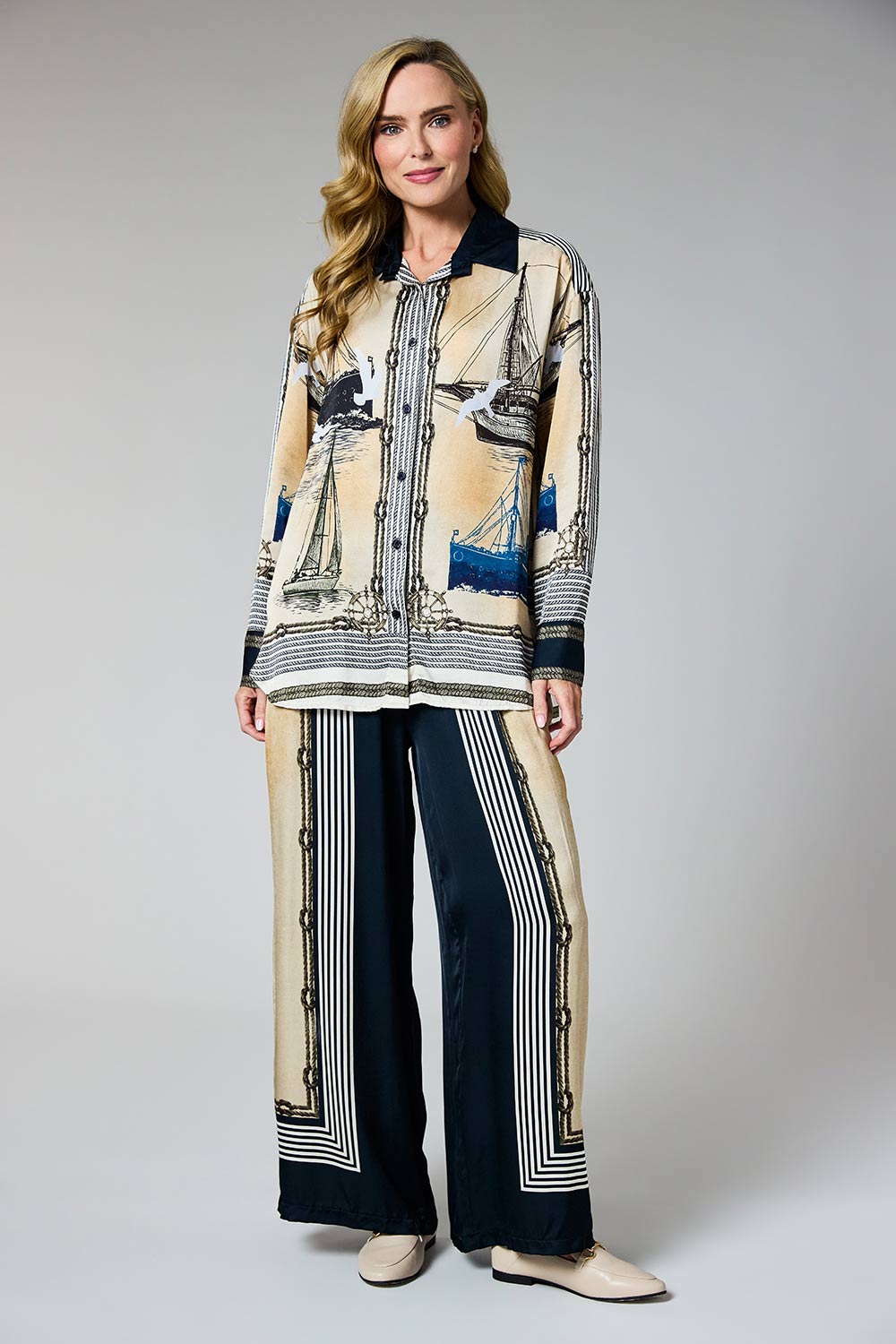 A woman modeling a matching two-piece outfit consisting of a beige and navy blue printed button-up shirt with a nautical boat pattern, and matching wide-leg trousers with vertical navy and beige stripes.
