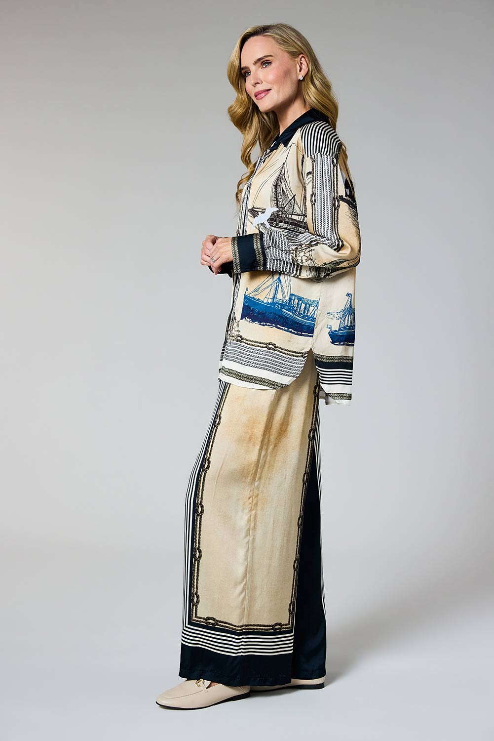 A woman models a long-sleeved cream and navy blue patterned jacket with matching wide-leg trousers. The jacket features a nautical design with illustrations of sailboats and stripes, paired with solid navy cuffs.