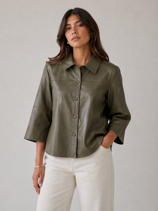 A woman modeling a cropped, olive green faux-leather button-up shirt with three-quarter sleeves, paired with off-white pants, against a neutral gray studio background.