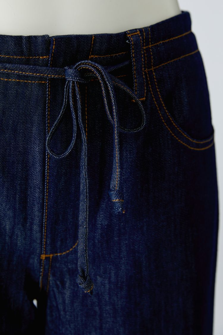 Close-up of dark indigo denim pants on a mannequin, showing the waistband with a matching drawstring, contrast orange stitching, and front pocket details.