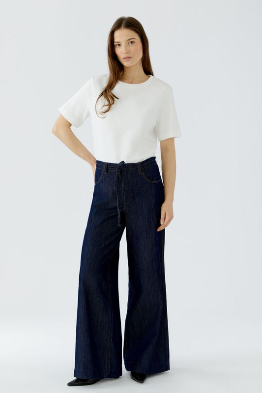 A woman modeling a white short-sleeve top and dark blue wide-leg denim jeans against a light gray studio background.