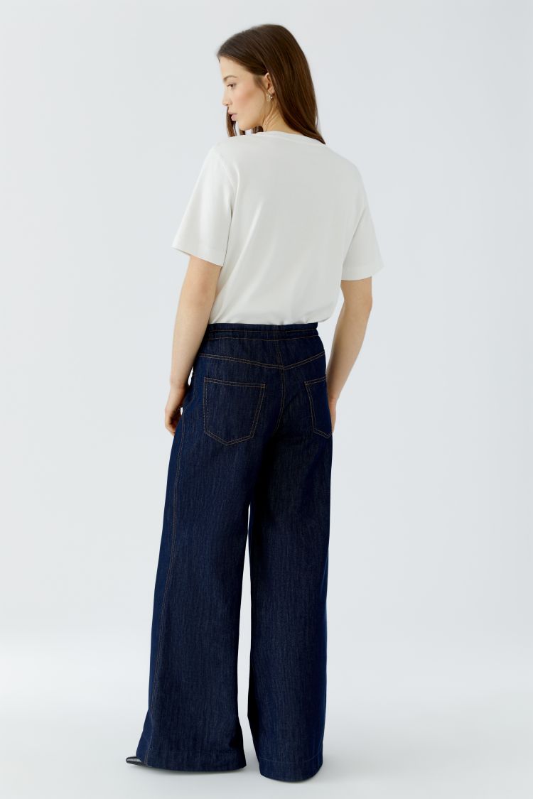A woman is shown from the back wearing dark blue wide-leg denim pants and a white short-sleeve t-shirt against a plain light gray studio background.