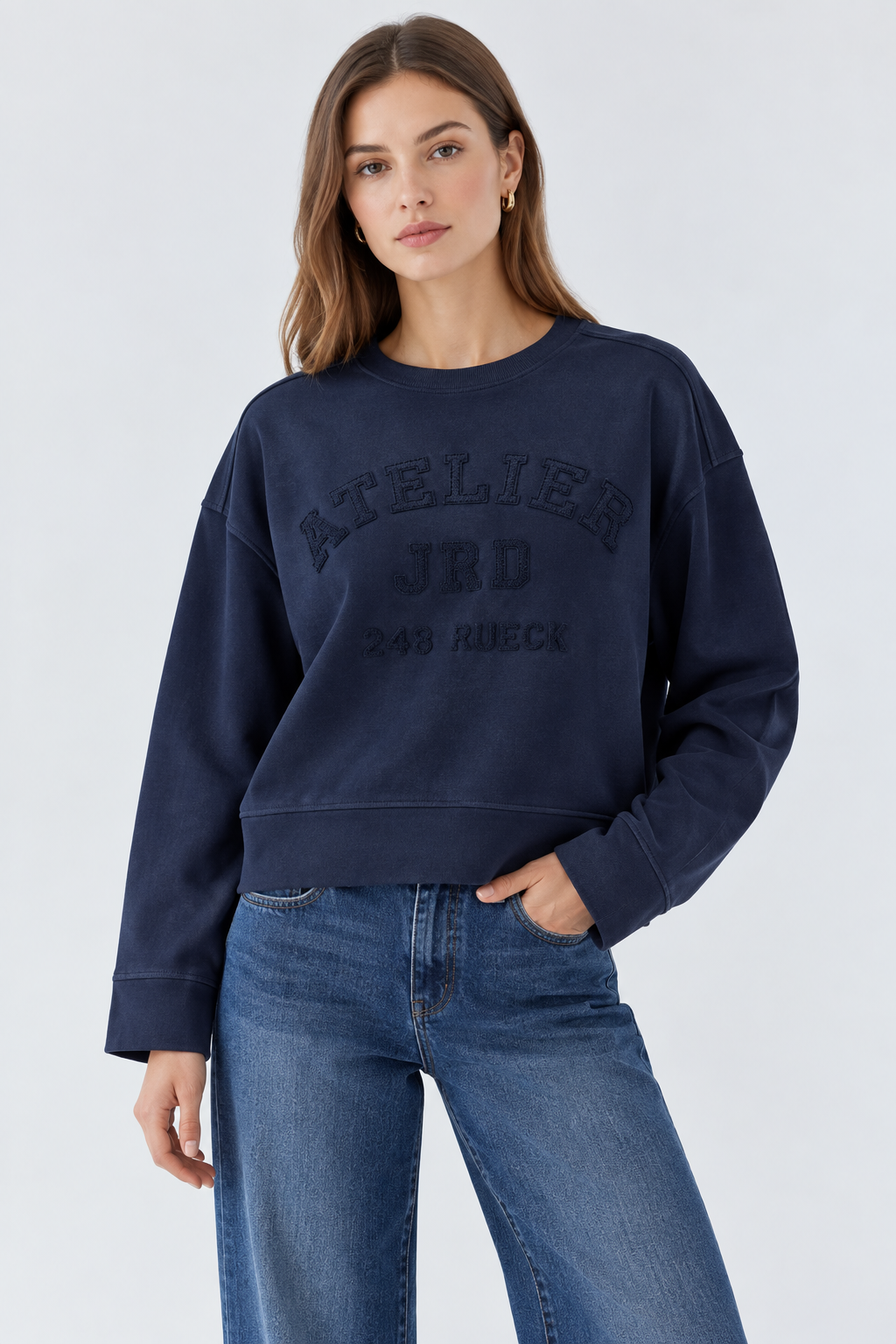A woman modeling a dark navy blue crewneck sweatshirt with embossed text reading 'ATELIER JRD 283 RIECK' on the front, paired with blue jeans against a light gray studio background.