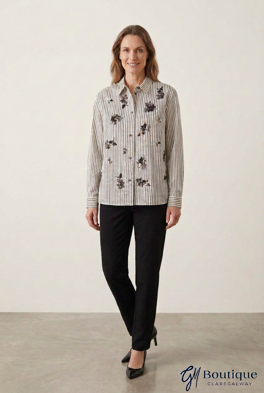 A woman modeling a long-sleeve button-up shirt with vertical black and white stripes and black floral embroidery, paired with black trousers and black heels, against a neutral studio background.