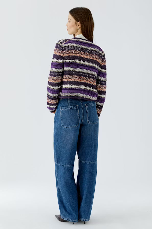 A woman shown from the back wearing a purple, black, and beige striped knit cardigan with wide-leg blue jeans, photographed against a light gray studio background.