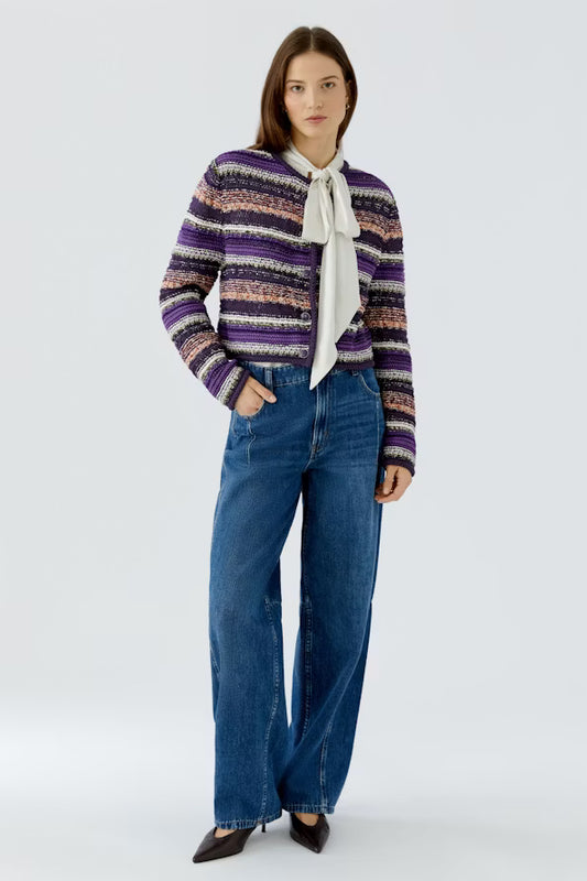 A model stands against a light gray studio background wearing a purple, black, and beige striped knit cardigan over a white bow-neck blouse, paired with wide-leg blue jeans and dark pointed flats.