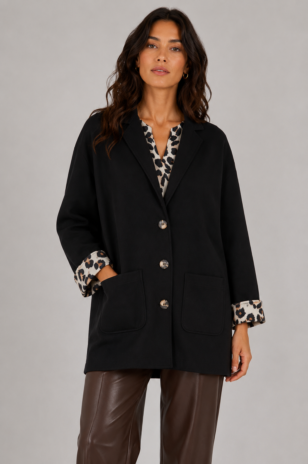 A woman modeling a black blazer with leopard-print patterned cuffs and a matching leopard-print shirt underneath, paired with brown pants, against a solid gray studio background.