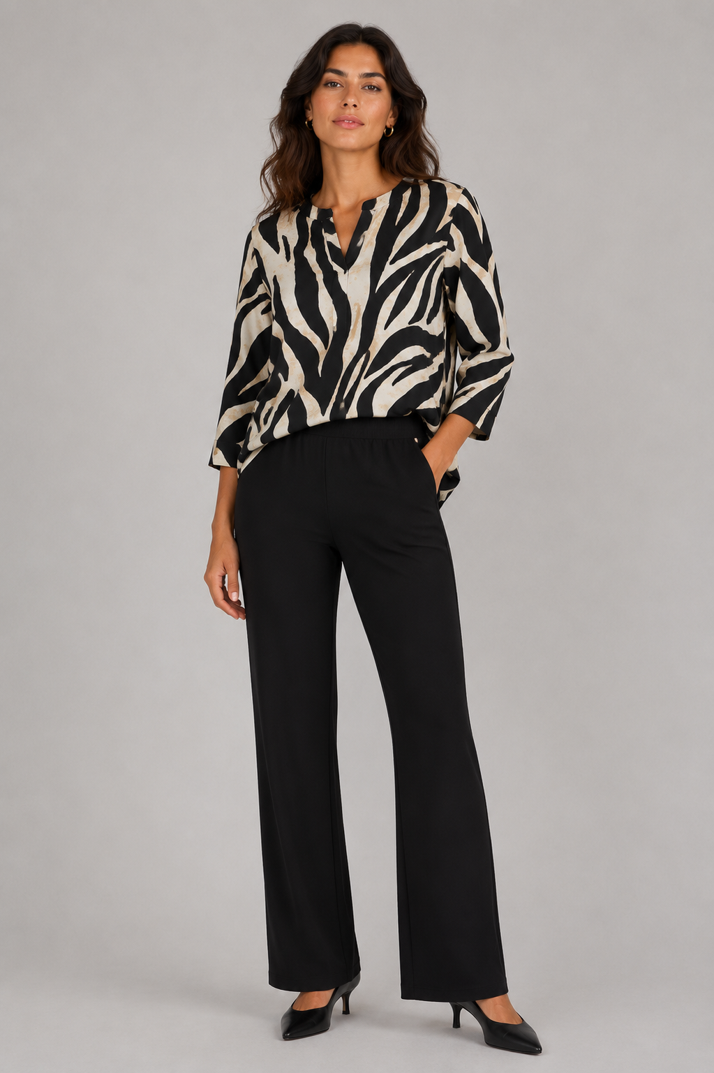 A woman modeling a black and beige zebra-print blouse with three-quarter sleeves, paired with black wide-leg trousers and black heels, against a solid gray studio background.